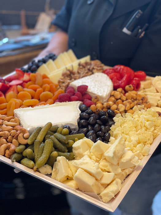 Image is a side view of the Signature Cheese Platter with olives, nuts, fresh and dried fruit and additional accoutrements being held by a BCS staff member. 