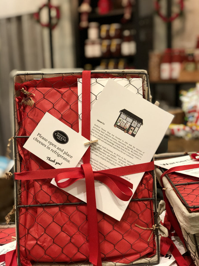 Image is of a holiday themed closed farmhouse style wire basket on its side. Finished with red tissue paper, ribbon, and Bedford Cheese Shop postcard.