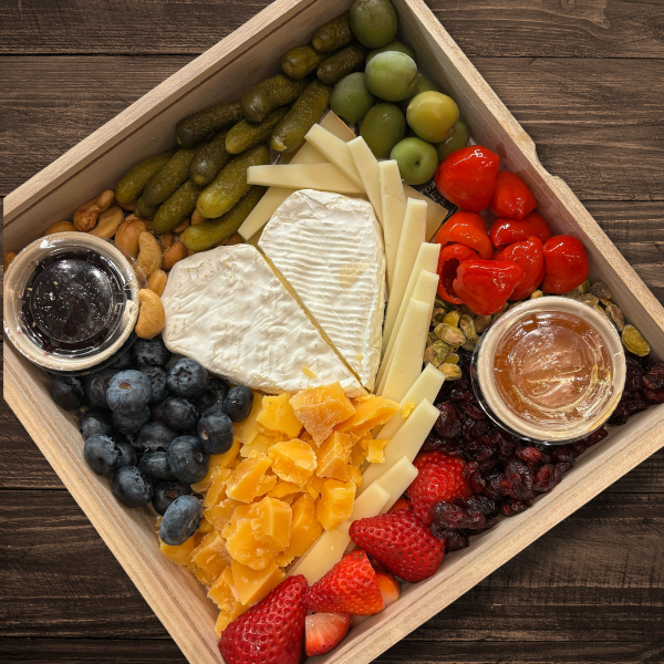 Image is of a cheese board to-go, displaying truncated cheese, wedged cheese, fresh seasonal fruit, nuts, honey, and jam.