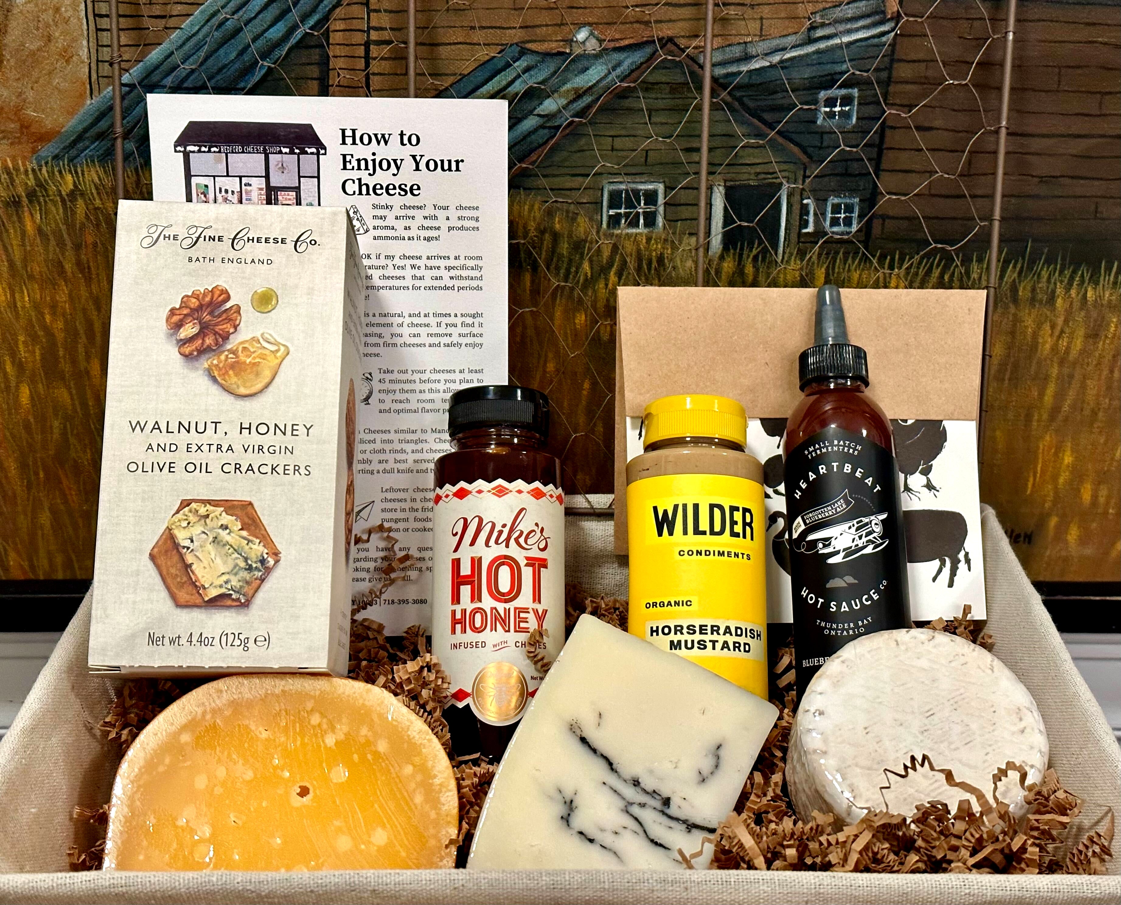 Image of the Washington Square Park gift basket contents inside a wire basket. Includes: hot honey, mustard, hot sauce, crackers, assorted cheeses, and a card.