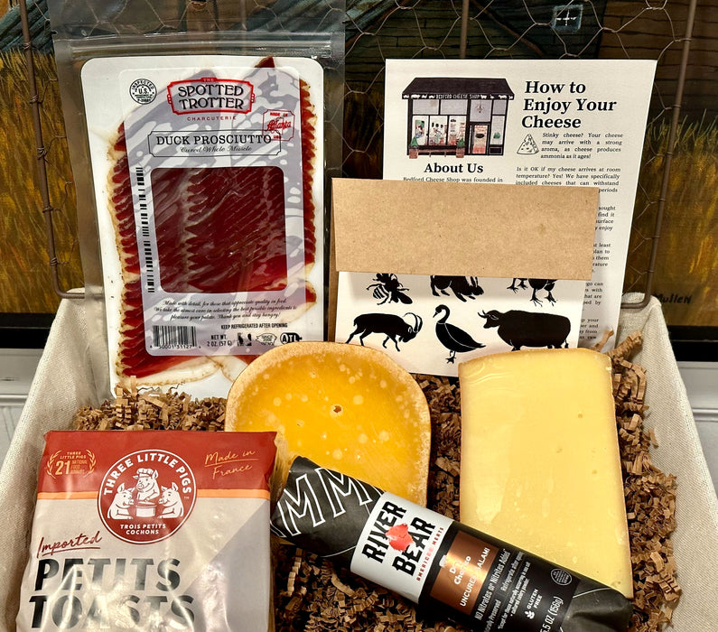 Image is of the Union Square gift basket contents displayed inside a wired basket. Including: duck prosciutto, chubs, toast points, cheeses, and a card. 