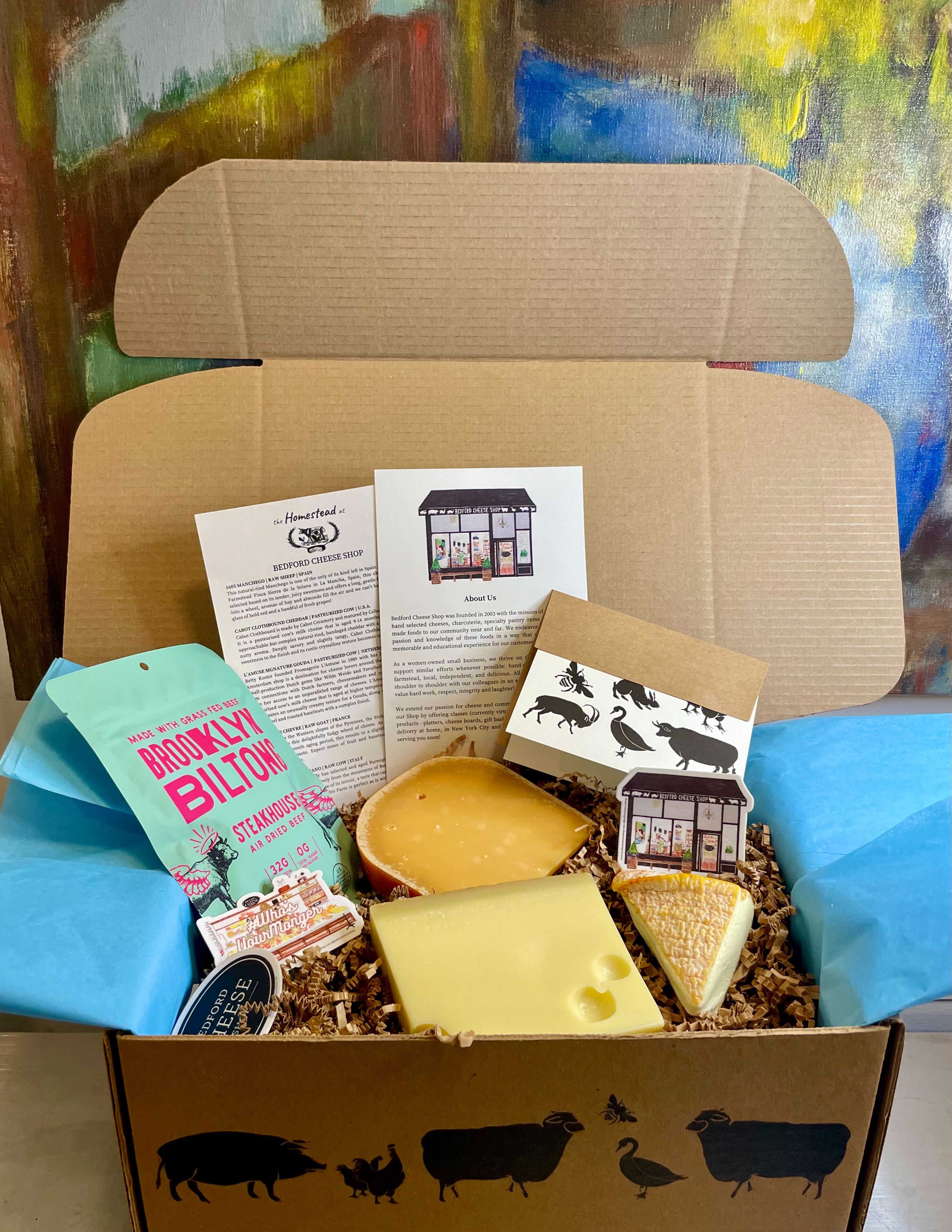 Image is of a monthly cheese fix subscription box. Depicted 3 hunks of cheese, jam, and BCS postcard nestled in brown crinkle paper inside of a BCS printed box.