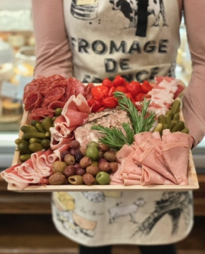 Image of a cheesemonger holding a charcuterie, meat only, board. Made up of thinly sliced cured meats, assorted olives, and pickles.Platters
