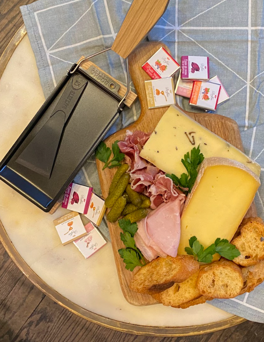 Image is of the raclette kit for two: a small raclette machine, cheese wedges, sliced meats, cornichons, and chocolate.