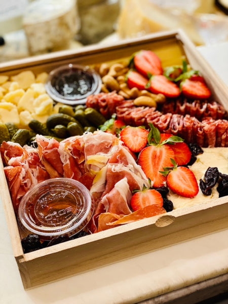 Image is of a cheese board to go with a charcuterie upgrade. Fresh halved strawberries, pickles, and thinly sliced cured meats surround the seasonal cheese selections on display in the wooden frame.