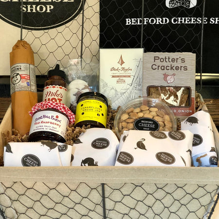 Image of The Original gift basket. A large sized  farmhouse style basket  full of assorted jams, honey, nuts, pickles, cheeses, cured meats, and sweet treats on a bed of cushy crinkle paper posed in front of the Bedford Cheese Shop logo.