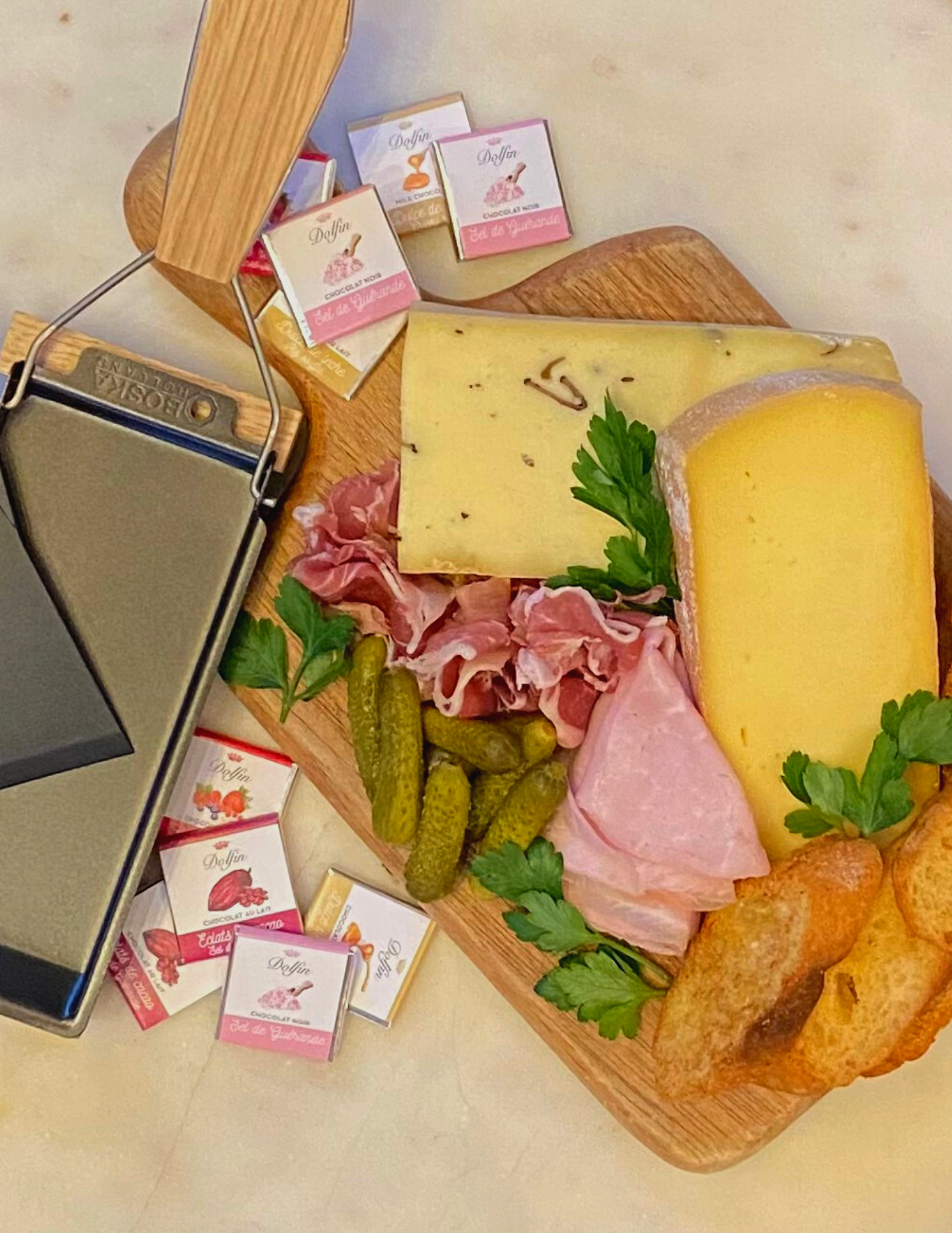 Image is of the raclette kit for two: a small raclette machine, cheese wedges, sliced meats, cornichons, and chocolate.