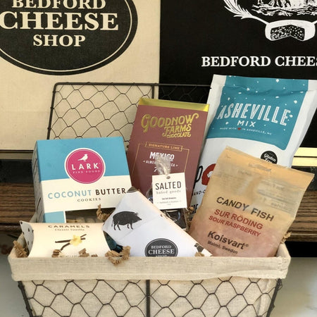 Image of The Sweets gift basket. A medium sized farmhouse style wire basket stuffed with candied popcorn, cookies, chocolates, assorted sweet treats and cheeses on a bed of cushy crinkle paper; posed in front of the Bedford Cheese Shop logo.