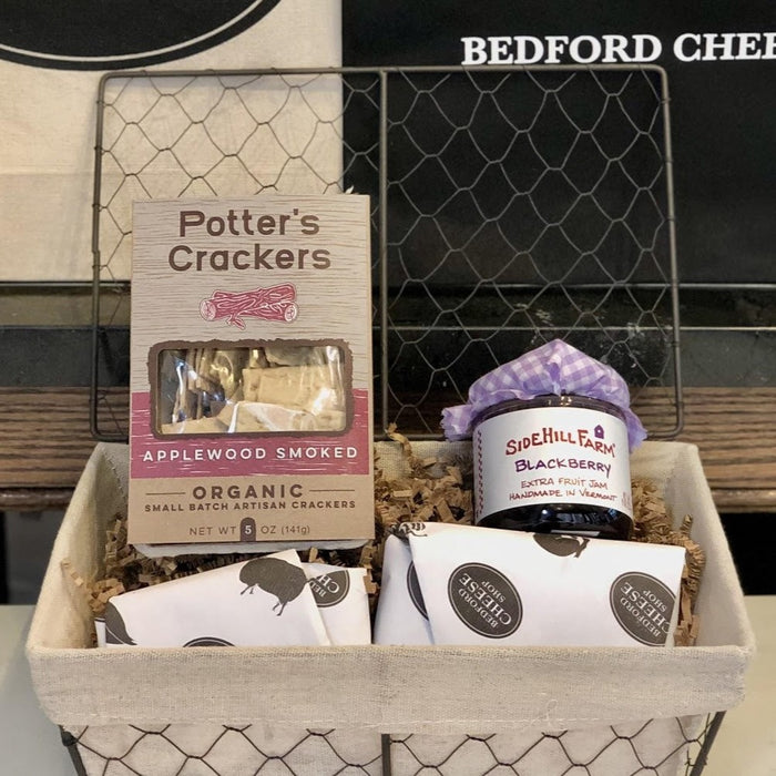 Image of The Hello gift basket. A small sized farmhouse style wire basket stuffed with crackers, a jam, and assorted cheeses on a bed of cushy crinkle paper; posed in front of the Bedford Cheese Shop logo.