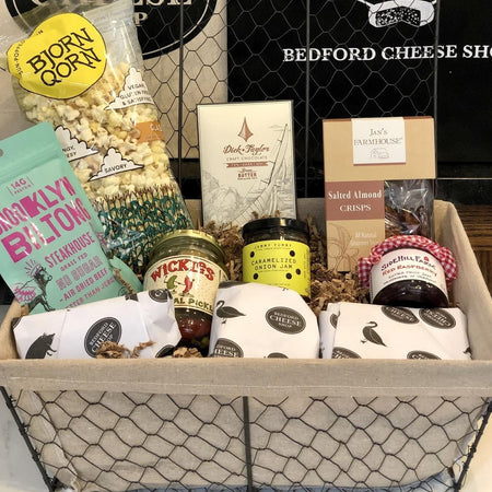 Image of the Simply the Best gift basket. A  large sized farmhouse style basket  full of assorted jams, pickles, cheeses, cured meats, and sweet treats on a bed of cushy crinkle paper posed in front of the Bedford Cheese Shop logo.